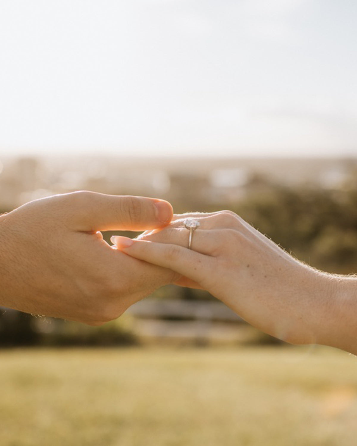 Couple holding hands outdoors, featuring a sparkling diamond engagement ring. Sunny background with blurred greenery.