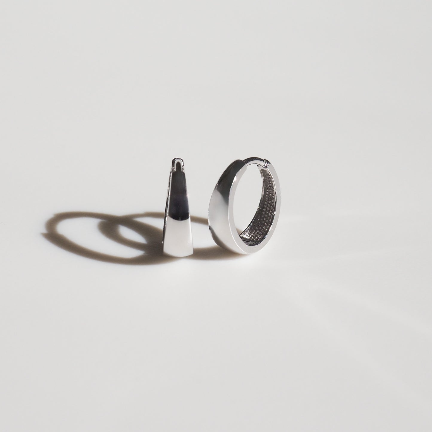 Minimalist silver earrings on a white background, featuring one hoop and one geometric design, casting subtle shadows. Luxurious jewellery.