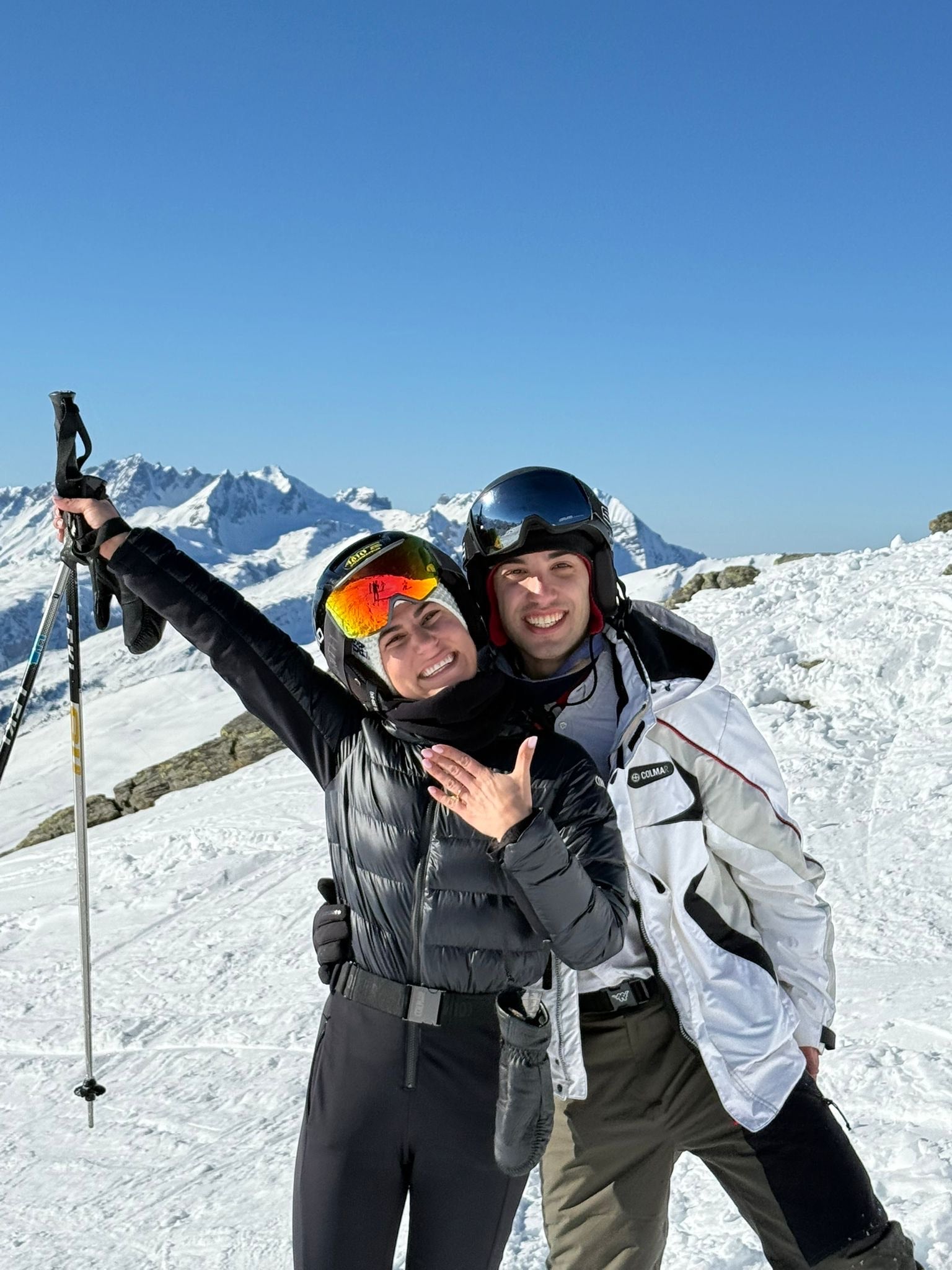 Couple in ski gear on snowy mountain, woman shows diamond ring with joy. Blue sky, sunny winter landscape. Luxury jewellery, engagement, travel.