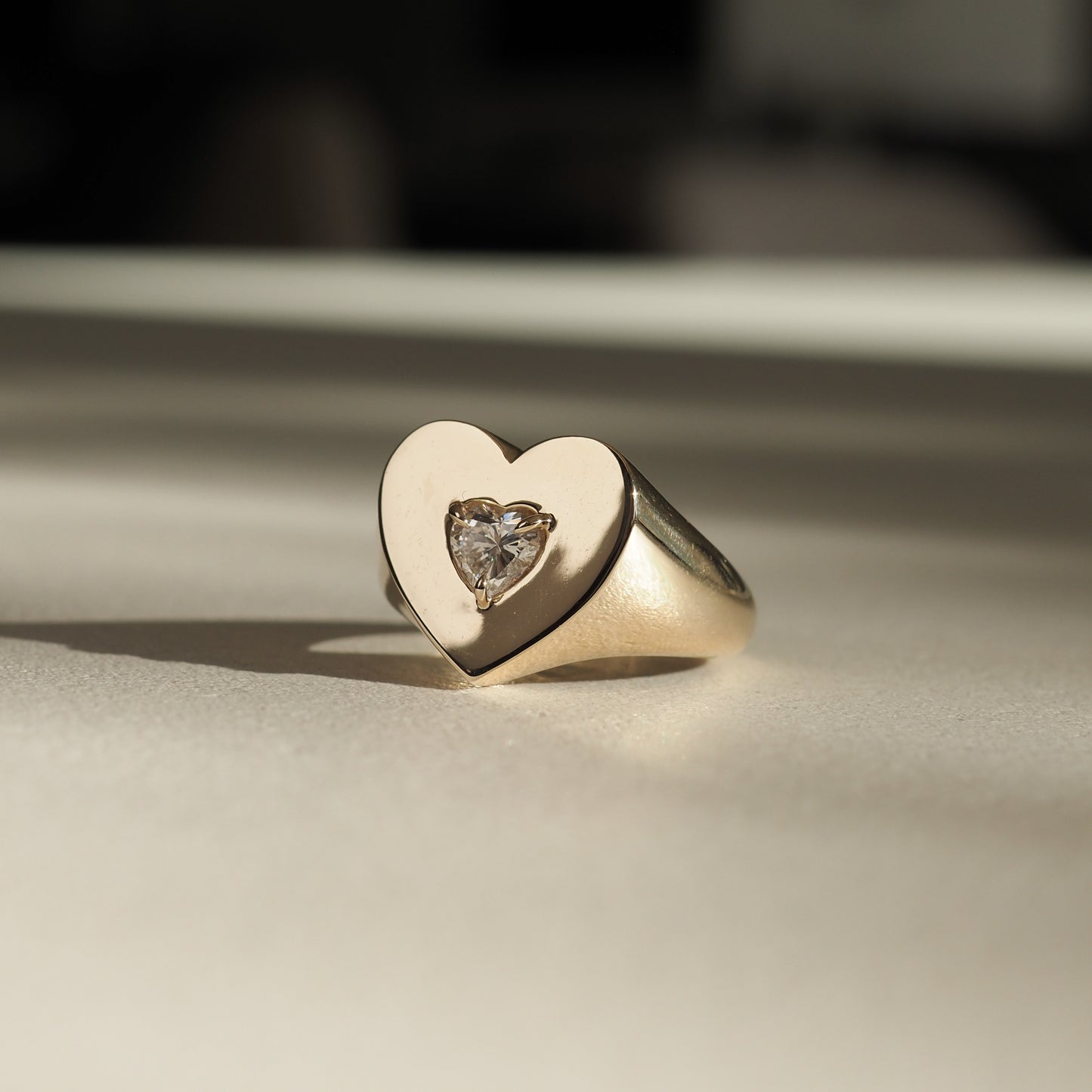 Gold heart-shaped ring with a central heart-cut diamond, resting on a smooth surface, showcasing luxury jewellery elegance.