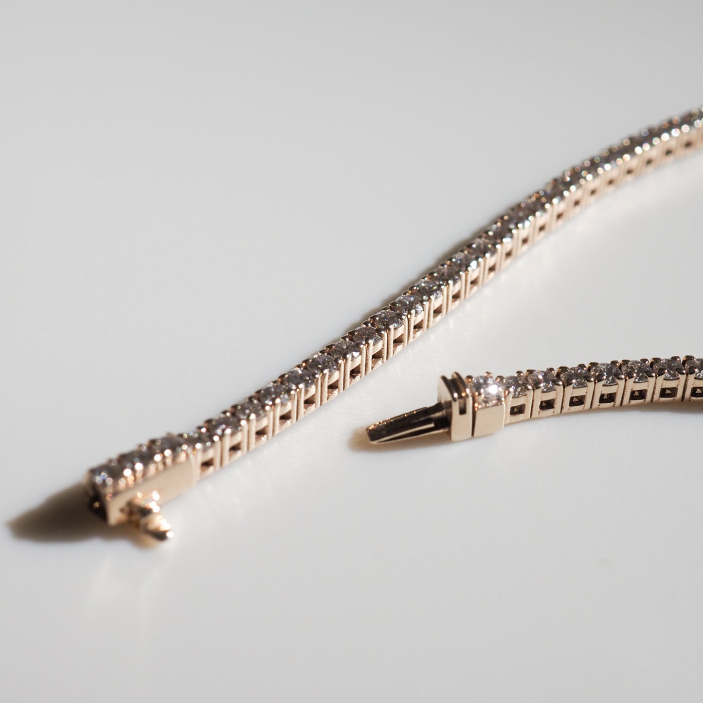Gold diamond tennis bracelet on a white surface, showcasing its clasp and shimmering stones, epitomising luxury jewellery craftsmanship.
