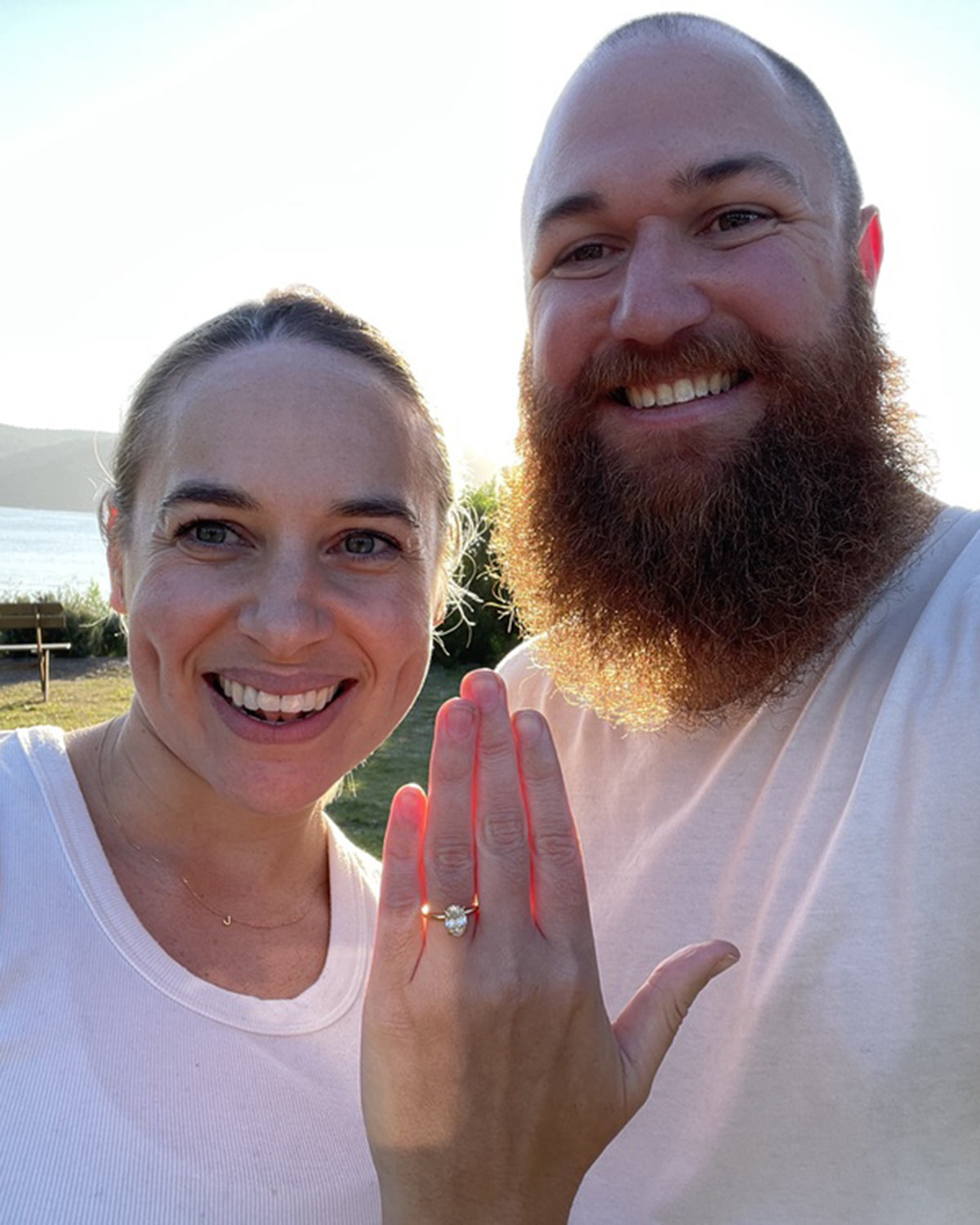Couple smiling outdoors; woman displays elegant diamond engagement ring. Sunlit background suggests a scenic, joyful moment.