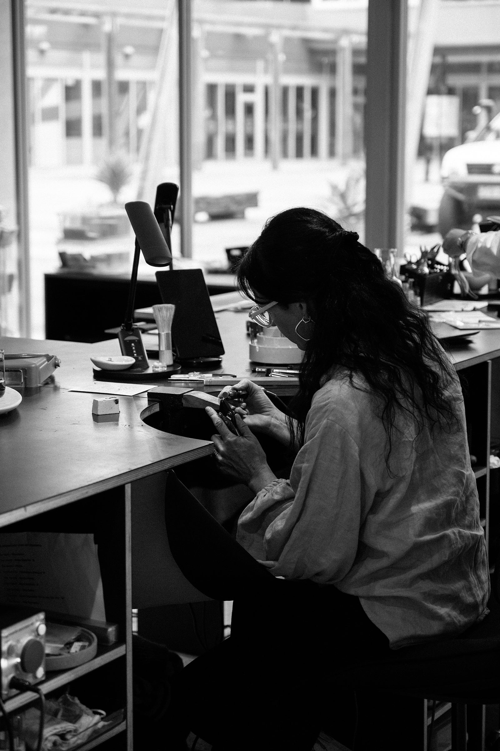 Jeweller crafting at workbench in a sunlit studio, surrounded by tools and materials. High-end jewellery making in progress. Black and white photo.