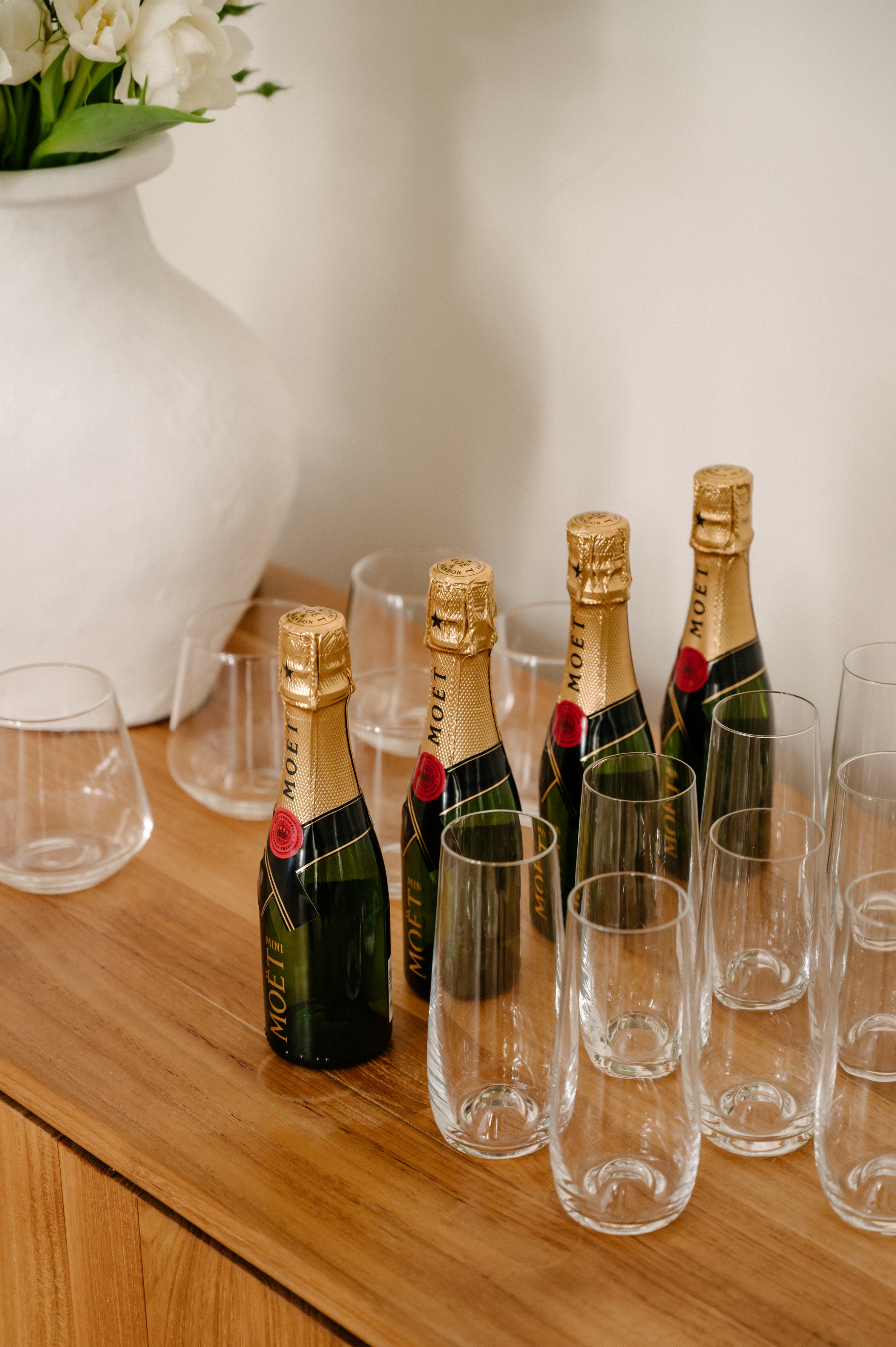 Champagne bottles and flutes on a wooden table beside a white vase with flowers, evoking luxury and celebration.