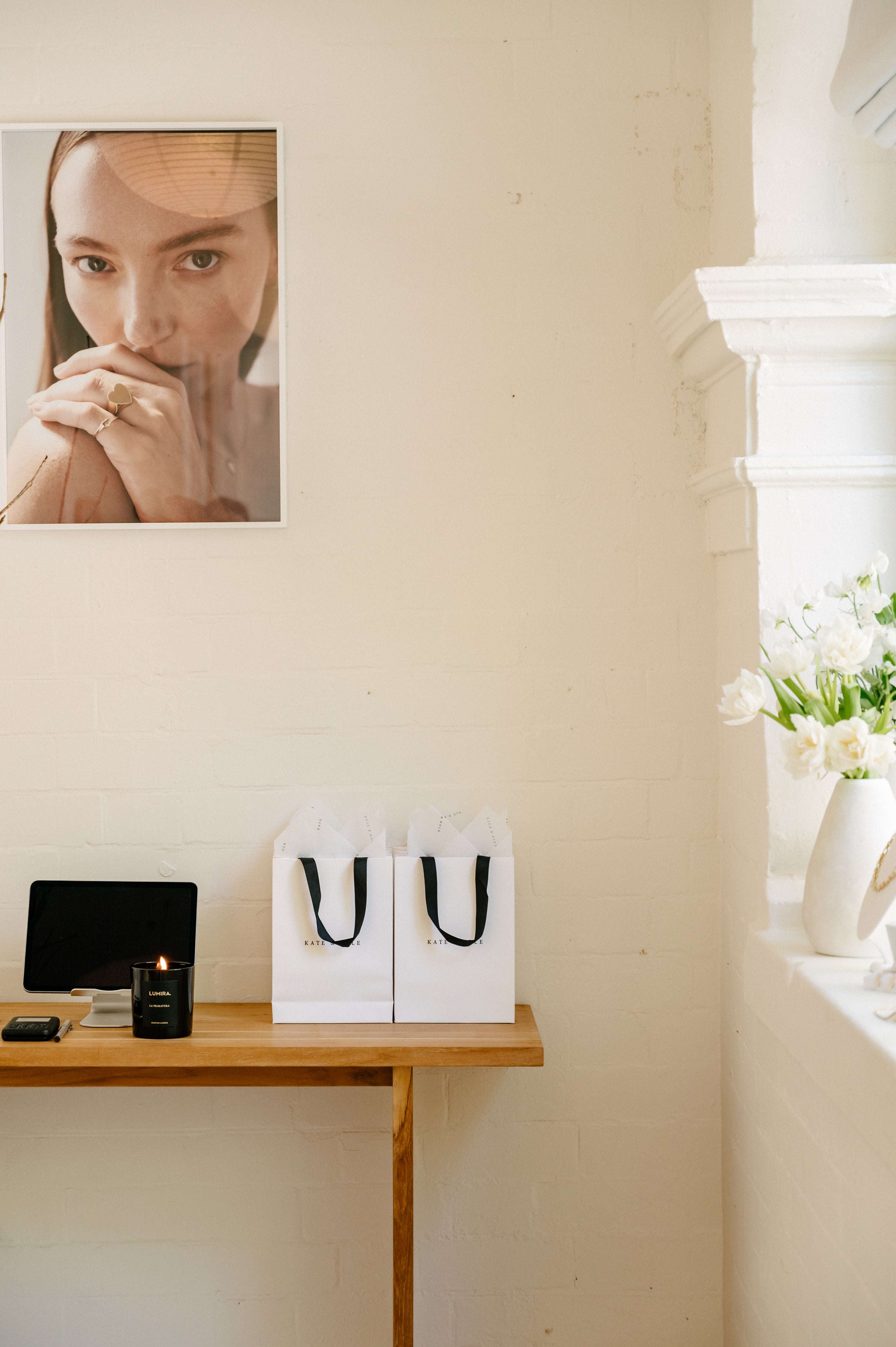 A minimalist room with a jewellery poster, two luxury gift bags on a wooden table, and white flowers in a vase by a window.