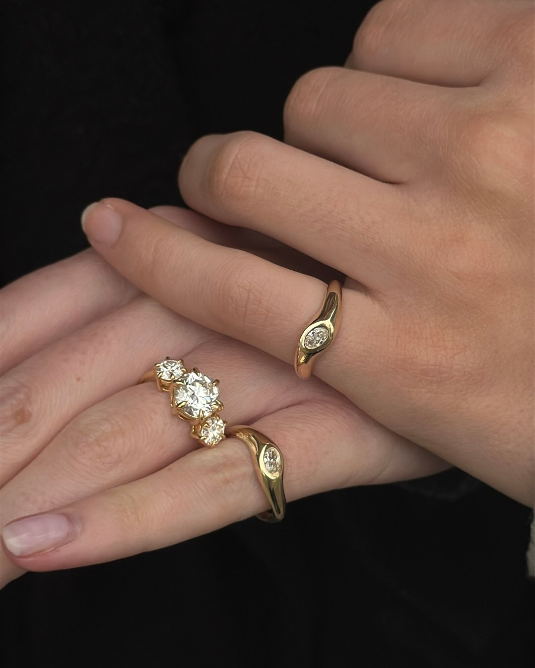 Gold rings worn on a person's fingers against a dark background, including a triple-stone diamond ring and a slim oval diamond ring.