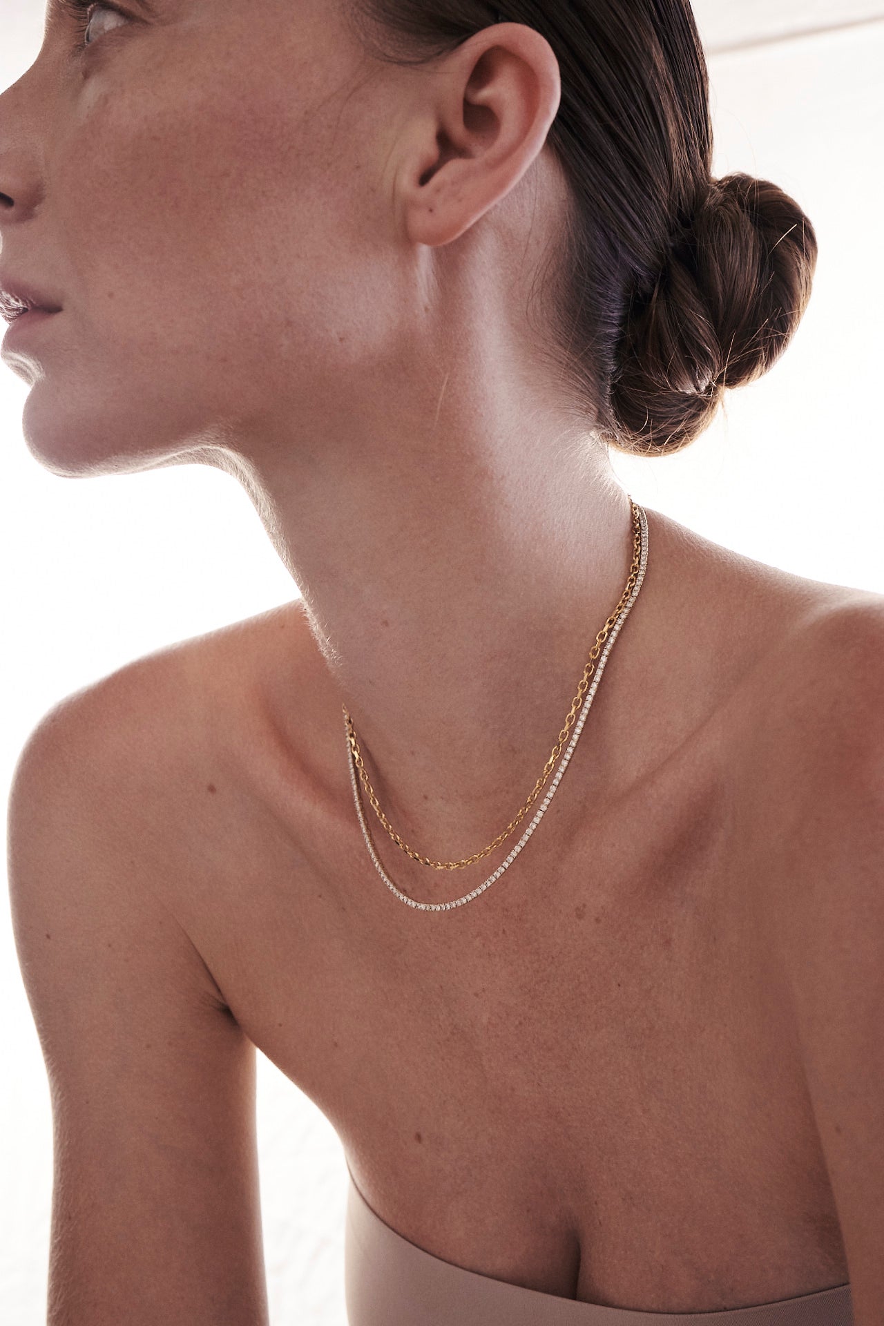 A woman with a sleek bun wears layered gold necklaces, showcasing luxury jewellery against a light background.