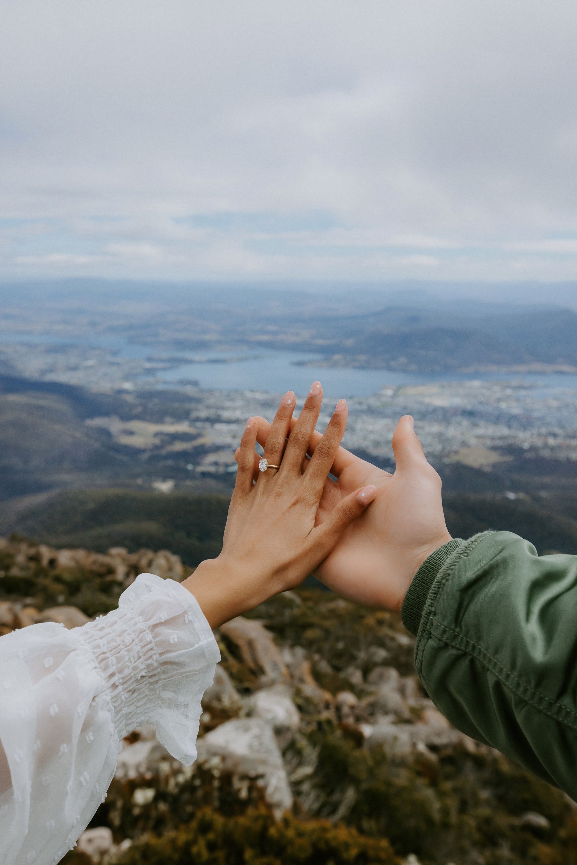 Couple holding hands with a diamond engagement ring, set against a panoramic mountain and valley view. Elegant jewellery, scenic backdrop.