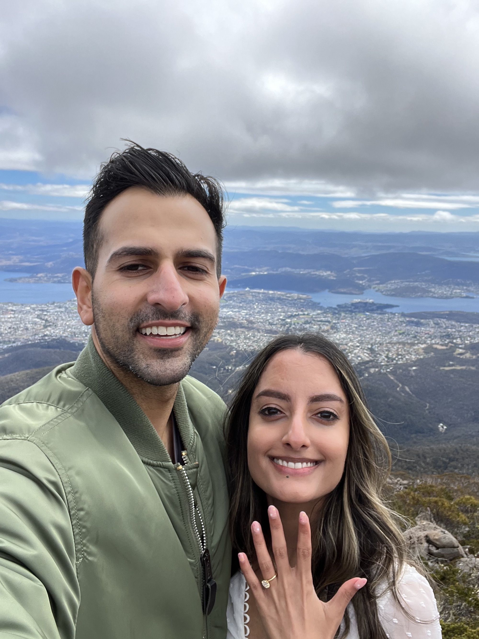 A couple smiles with a scenic mountain view, as the woman shows off an elegant engagement ring, symbolising love and commitment.