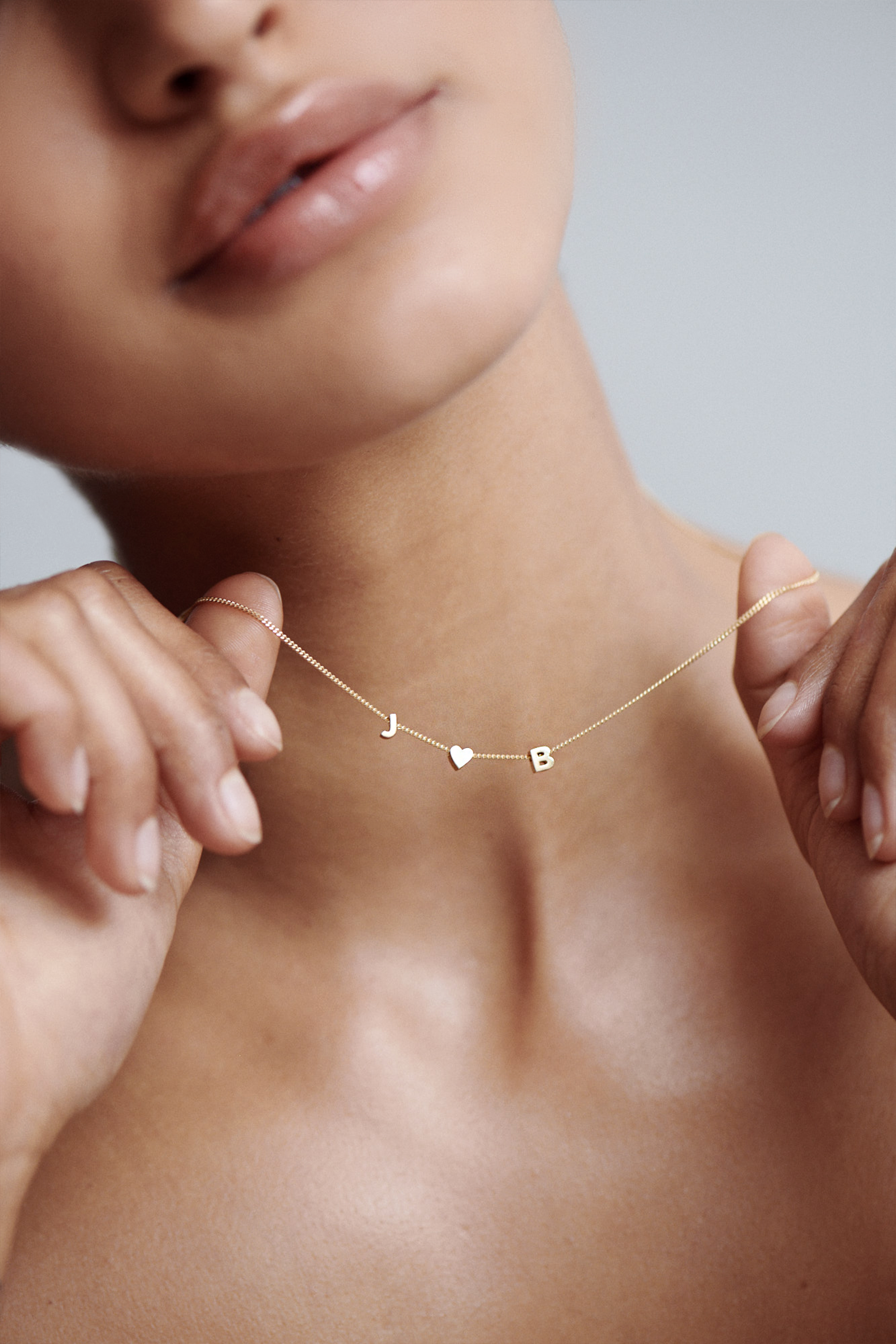 Woman holding a delicate gold necklace with "J", a heart, and "B" charms, showcasing luxury personalised jewellery.