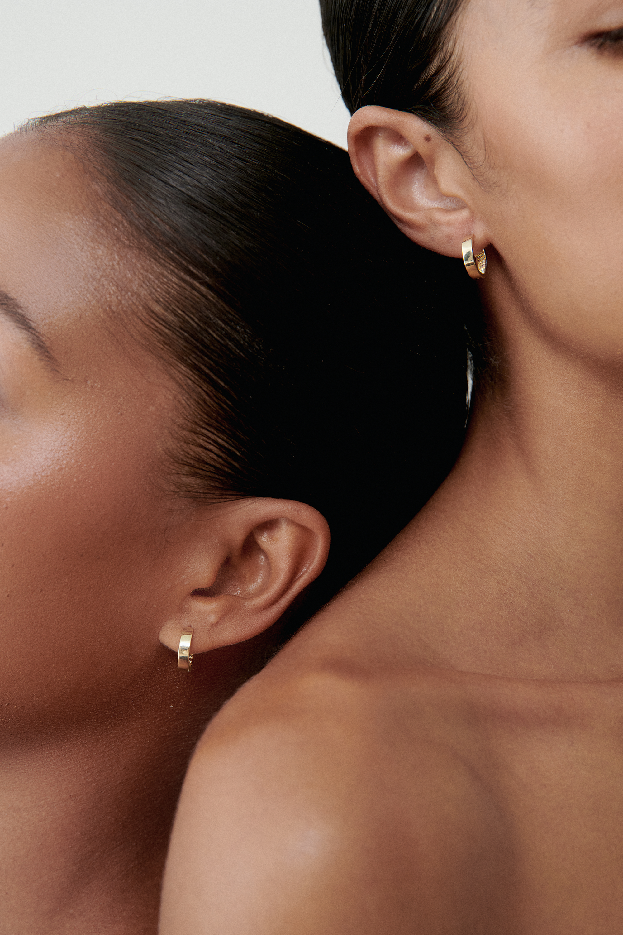Two women with sleek hairstyles showcasing elegant, minimalist gold hoop earrings. Luxury jewellery focus.