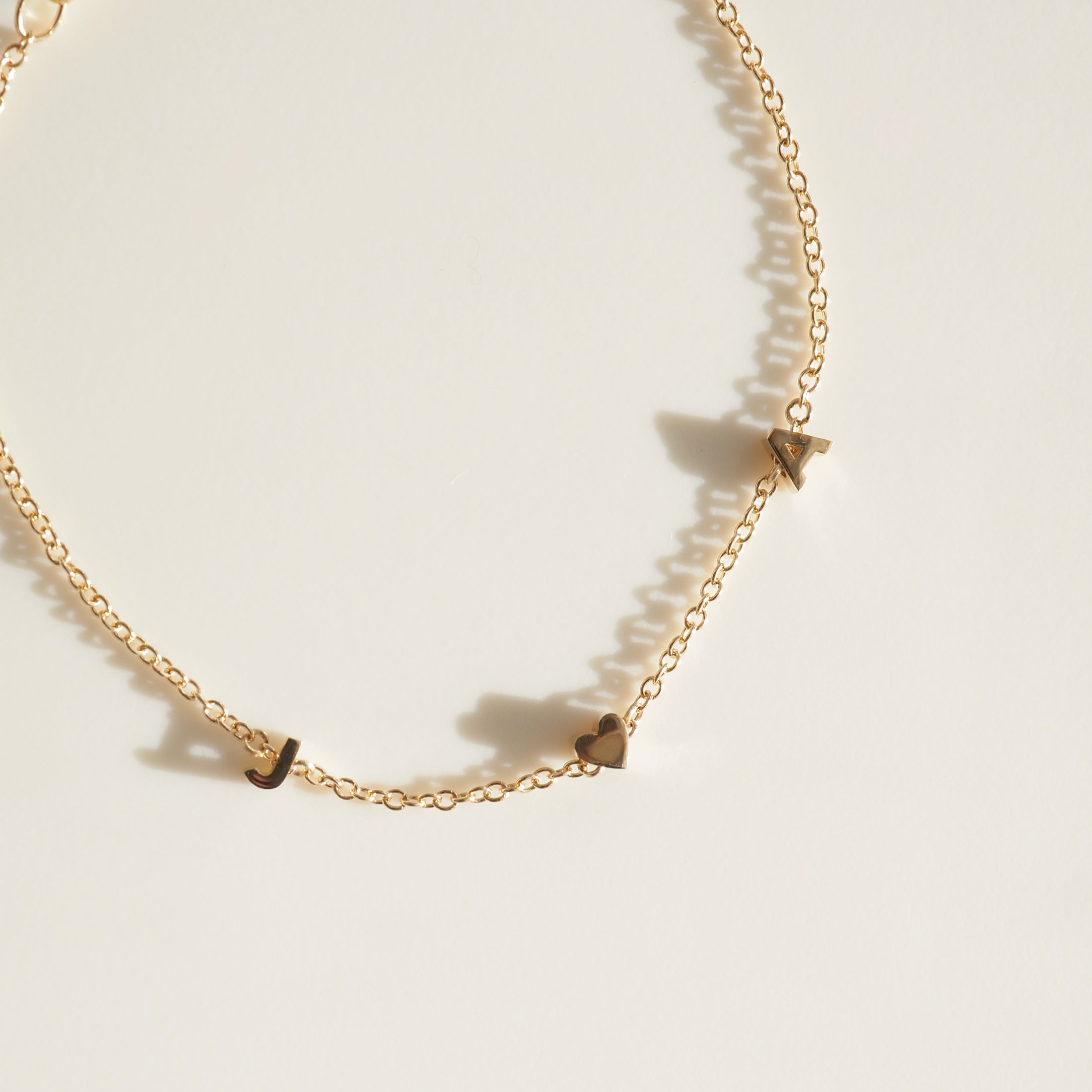 Gold necklace with delicate chain and charms: letter 'J', heart, and letter 'A'. Luxurious minimalist jewellery on a light background.