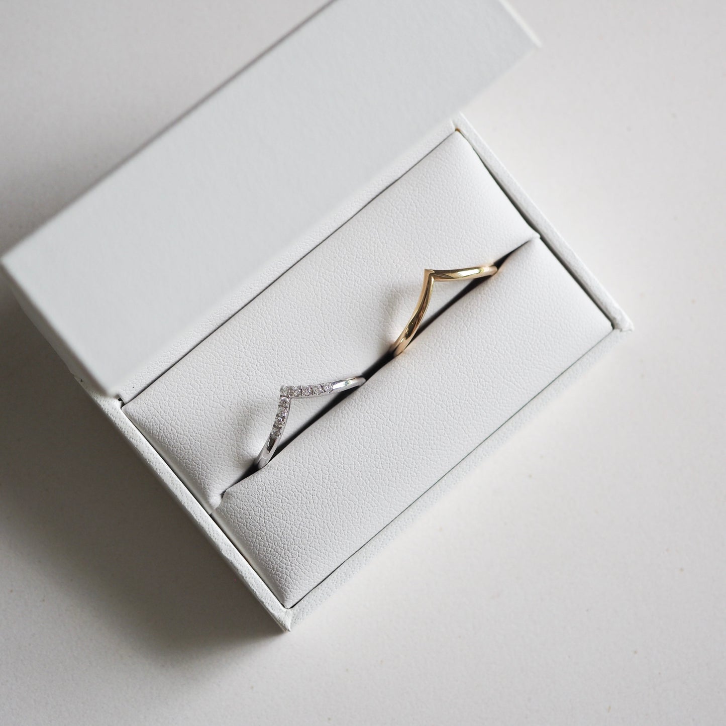 Elegant white jewellery box with two V-shaped rings; one silver with diamonds and one gold, set against a white backdrop.